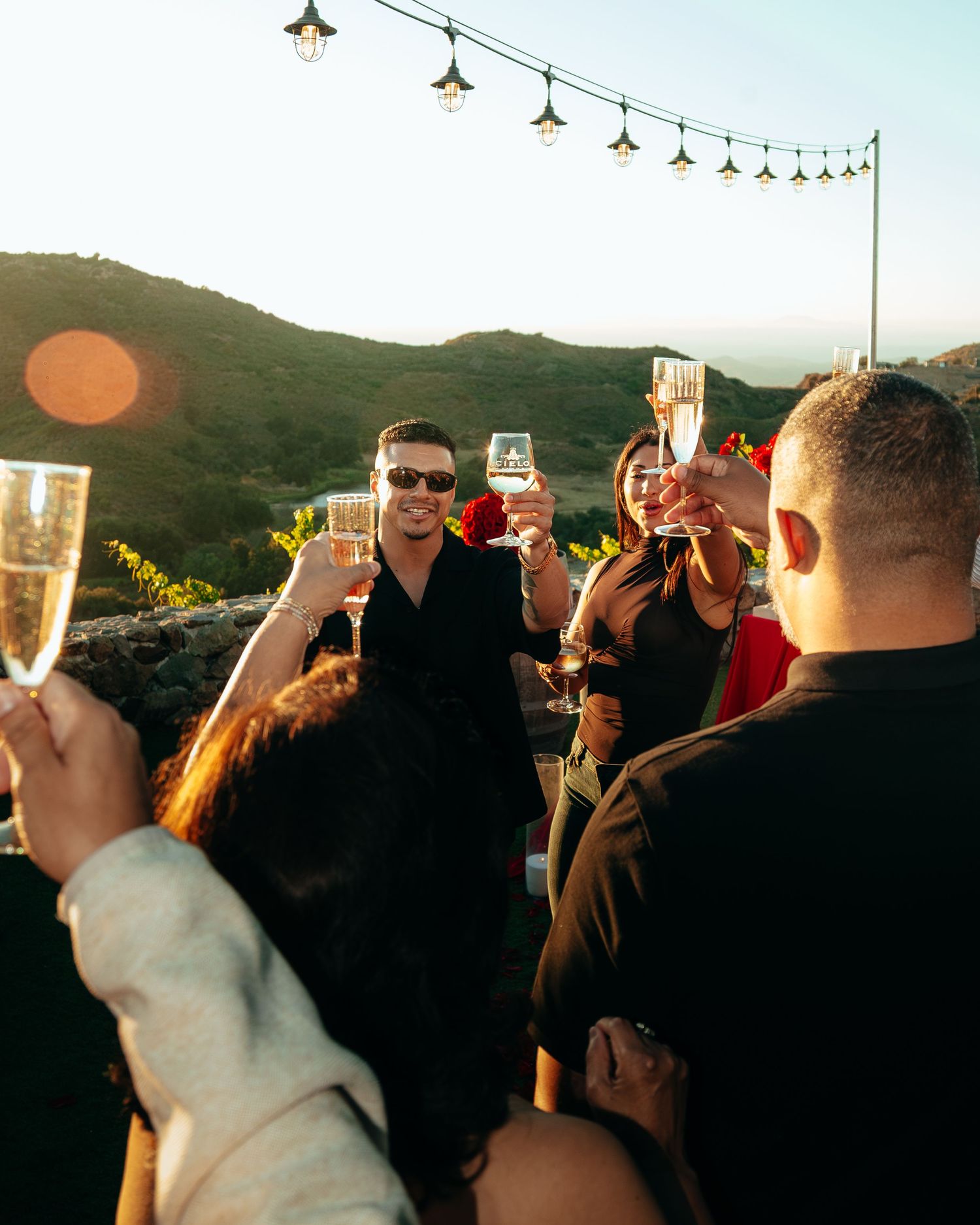 Group of friends toasting champagne glasses at golden hour outdoor celebration with scenic hillside backdrop.