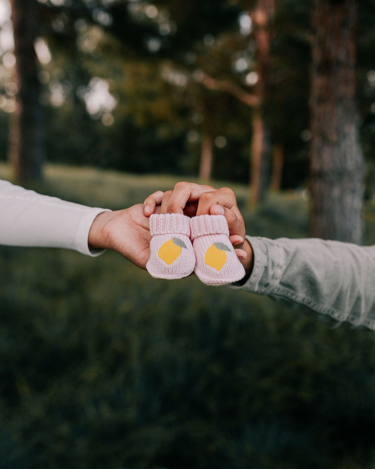 Two hands holding pink baby booties outdoors in forest setting.