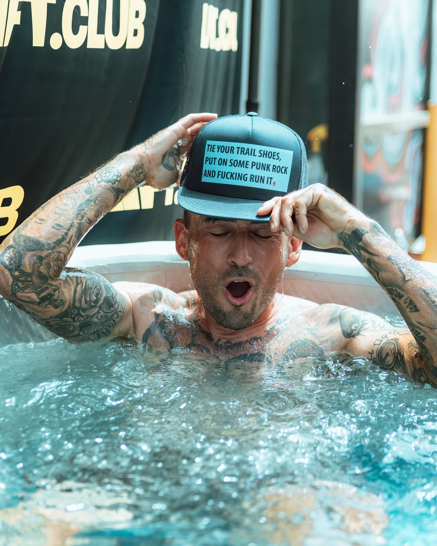 Heavily tattooed man in a trucker hat reacting while sitting in an ice bath at a fitness event.