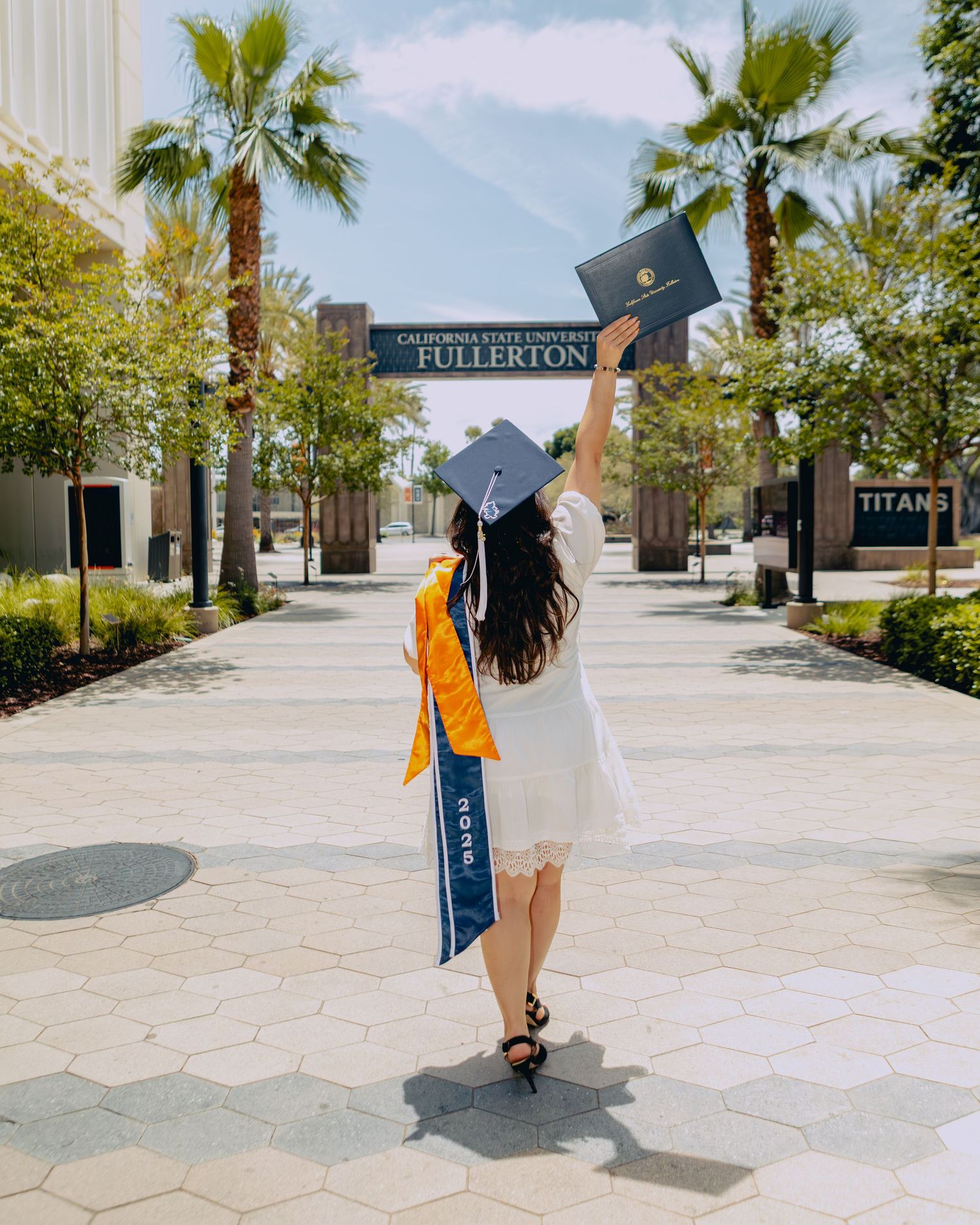 Graduate in white dress holds diploma aloft facing California State University Fullerton entrance sign.