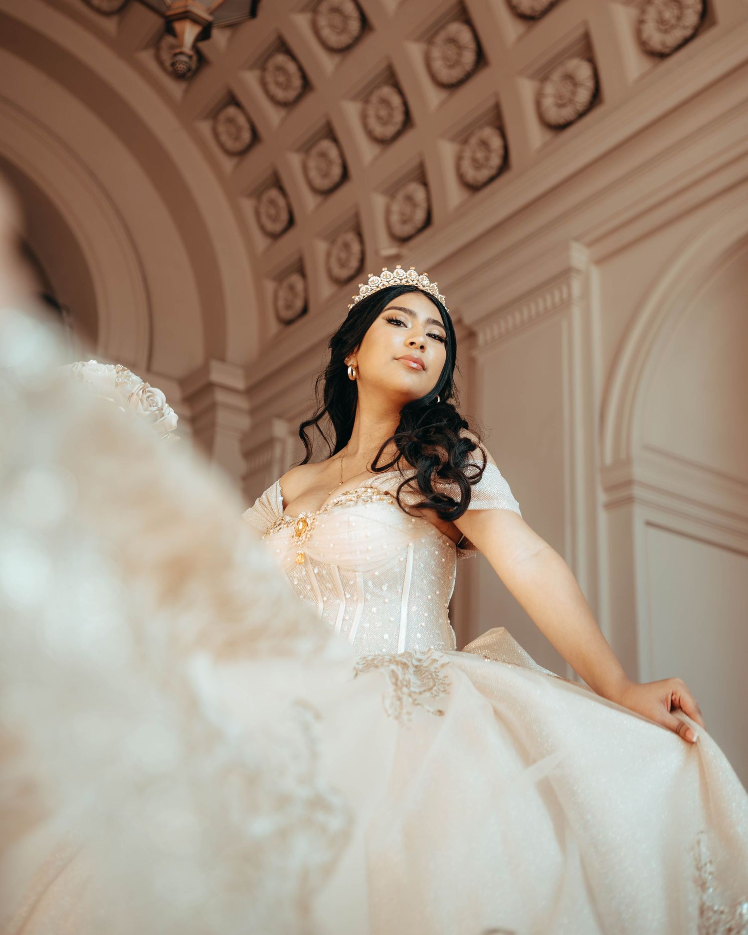 Elegant bride wearing a white ball gown and crown poses in an ornate ballroom.