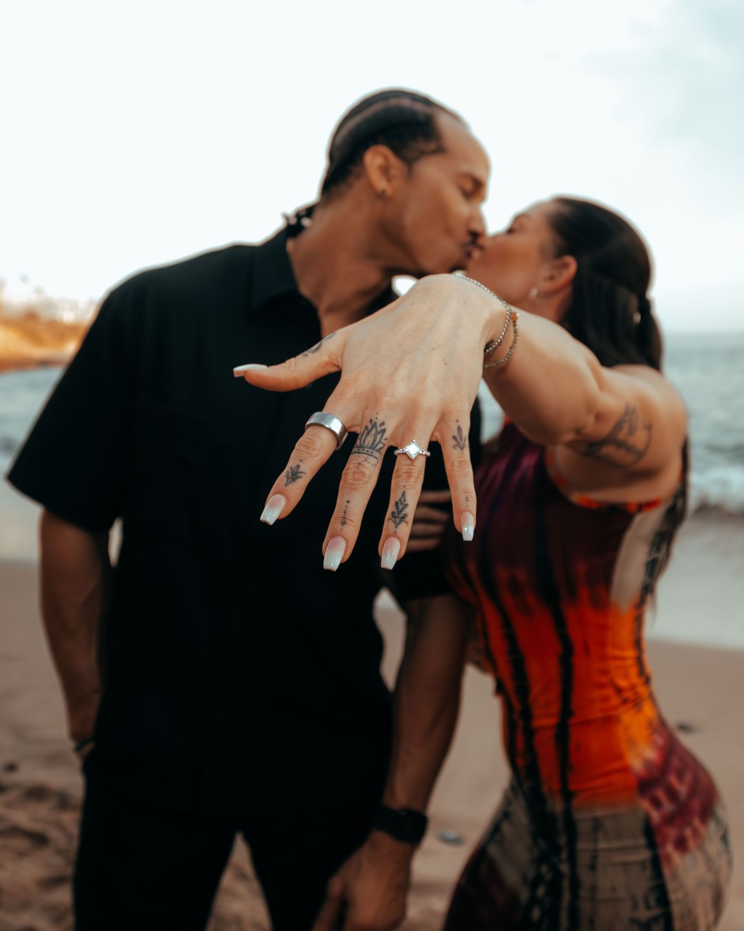 Couple kissing on beach while woman displays engagement ring with tattoos visible.