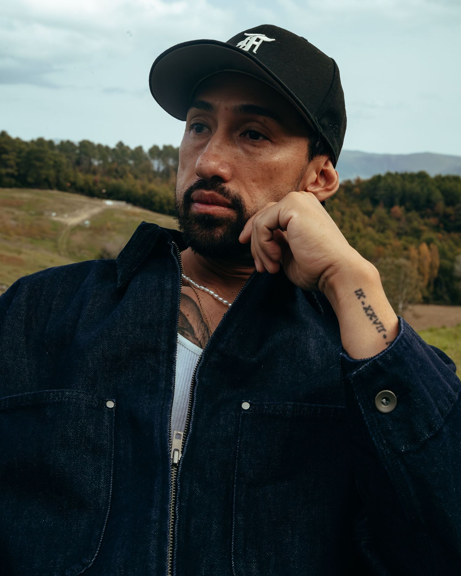 Man in black denim jacket and Art cap posing thoughtfully outdoors with autumn hills in background.