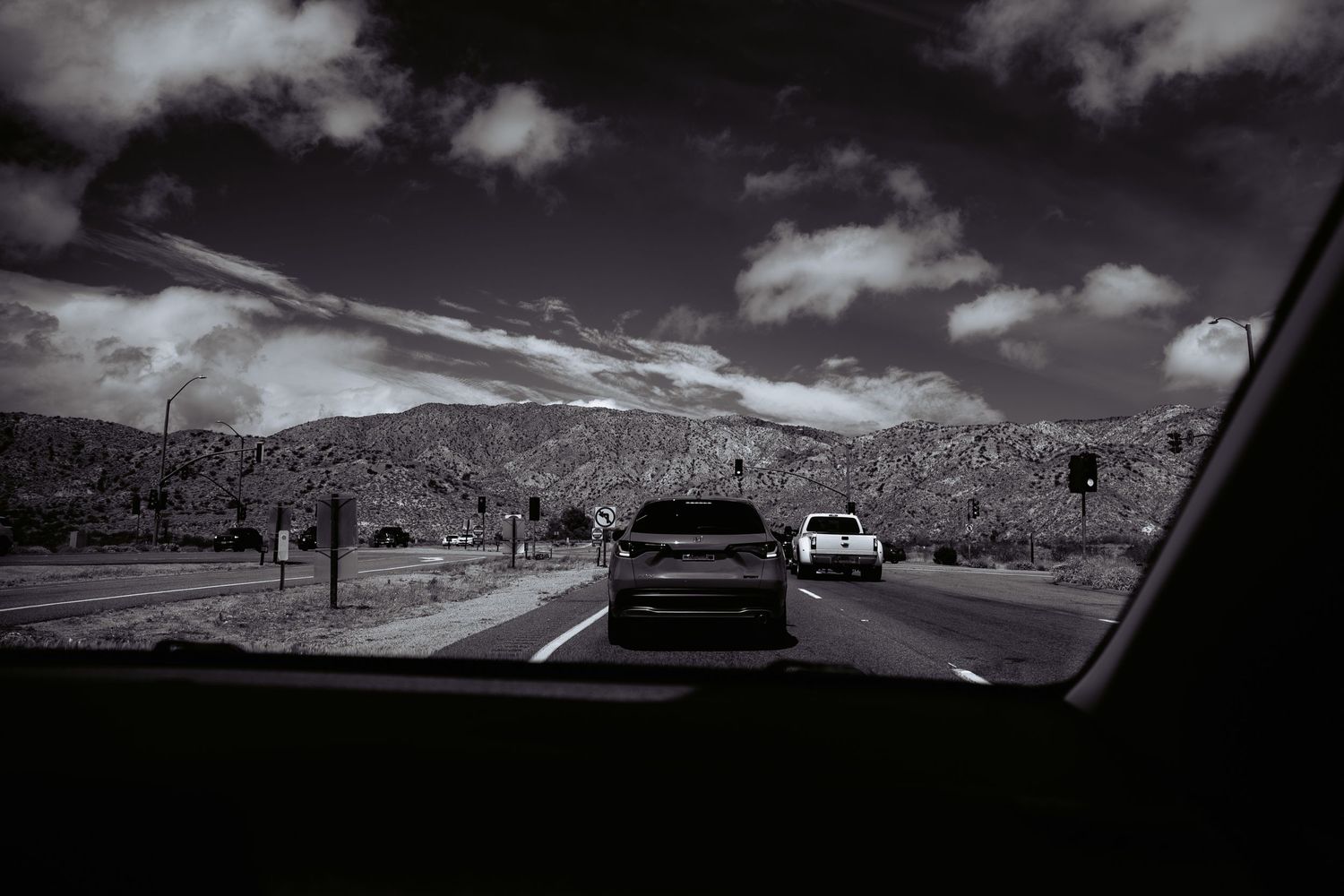 Black and white photo of cars driving on a desert highway with mountains and cloudy sky in background.