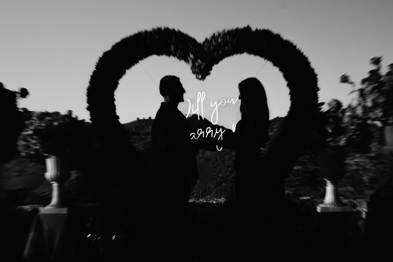 Romantic black and white silhouette of a couple standing beneath a heart-shaped floral arch at dusk.