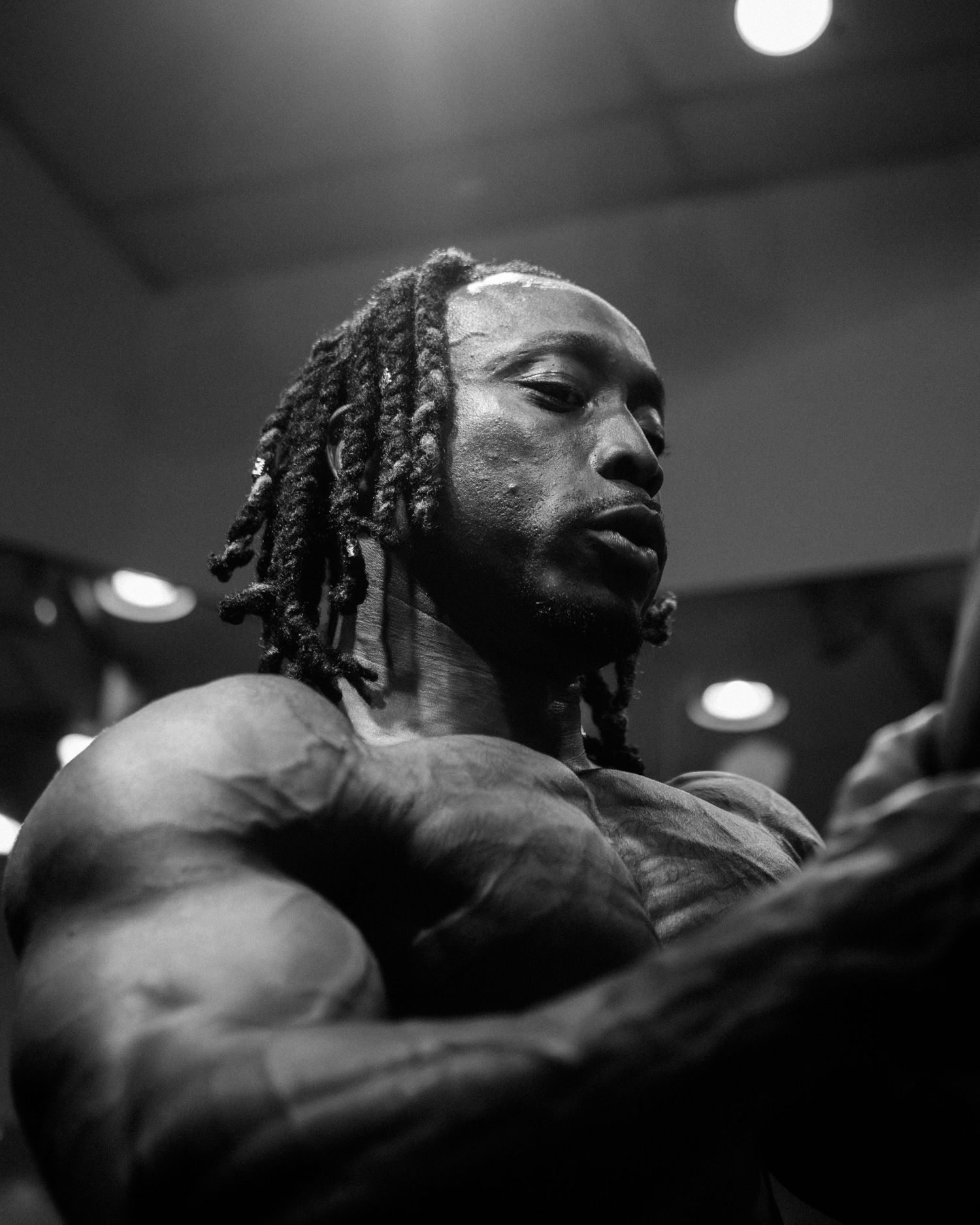 Black and white photo of a muscular bodybuilder with dreadlocks flexing under dramatic gym lighting.