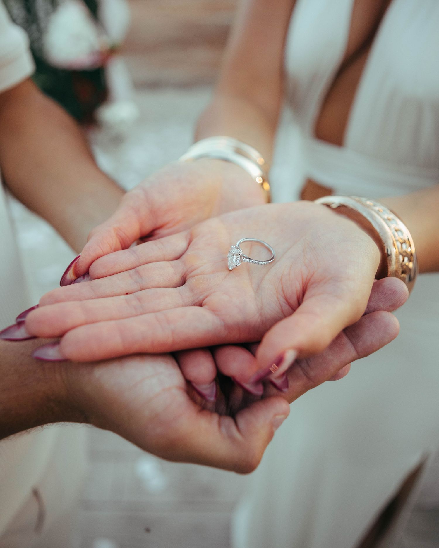 Hands cradling a diamond engagement ring with gold bracelet visible.