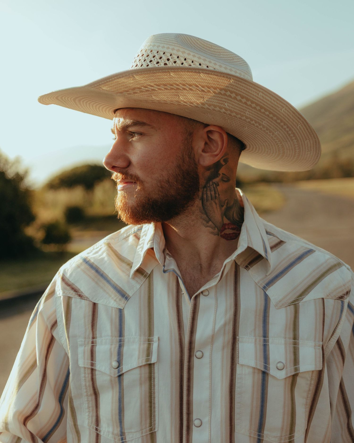 Bearded man with neck tattoo wearing cowboy hat and striped western shirt outdoors.