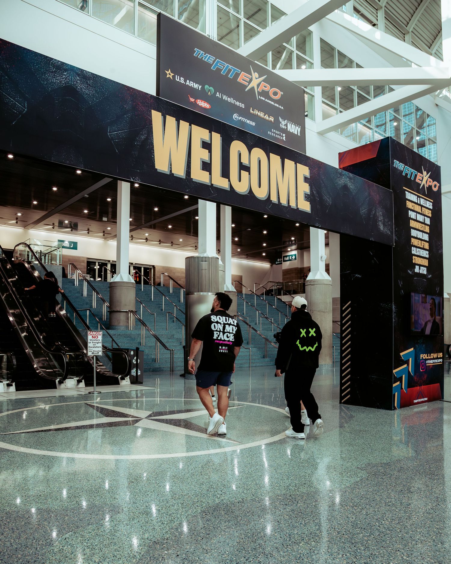 Two people walking toward a large Welcome sign at the Fit Expo convention center entrance.