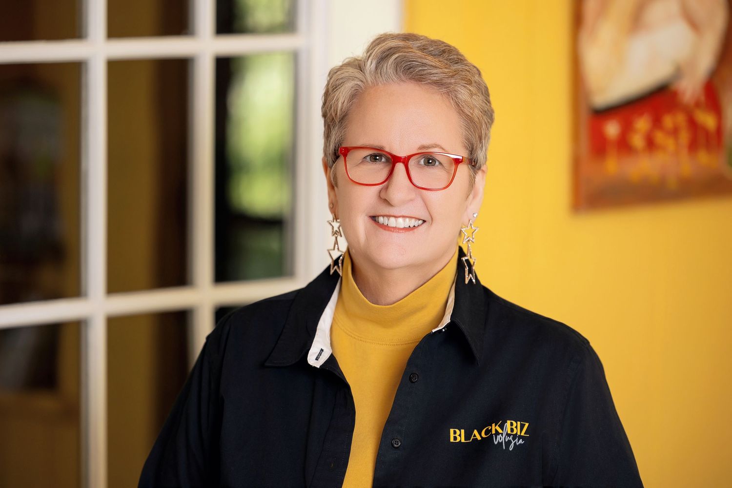Smiling woman with short gray hair wearing red glasses and a black jacket with yellow turtleneck.