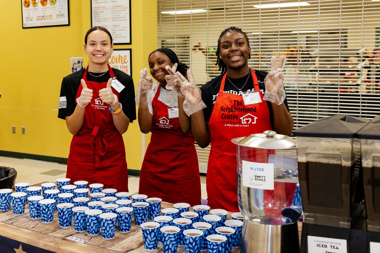 Three smiling volunteers in red aprons stand behind a table of blue cups at a community food service event.