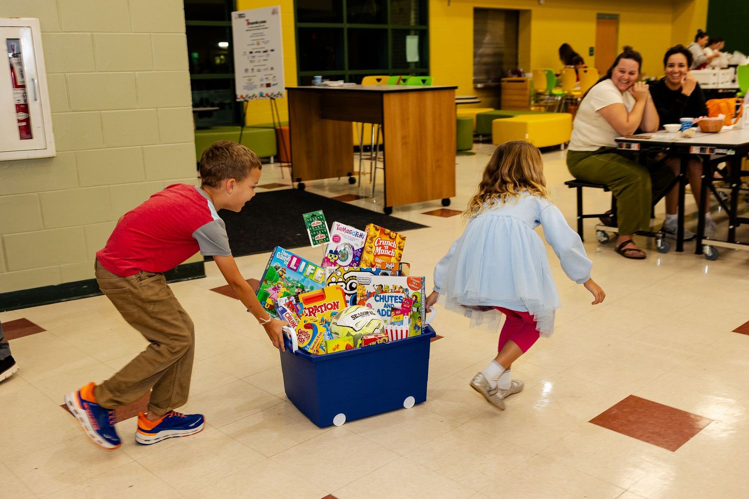 Two children playing and pushing a large bin filled with donated supplies in a school cafeteria.
