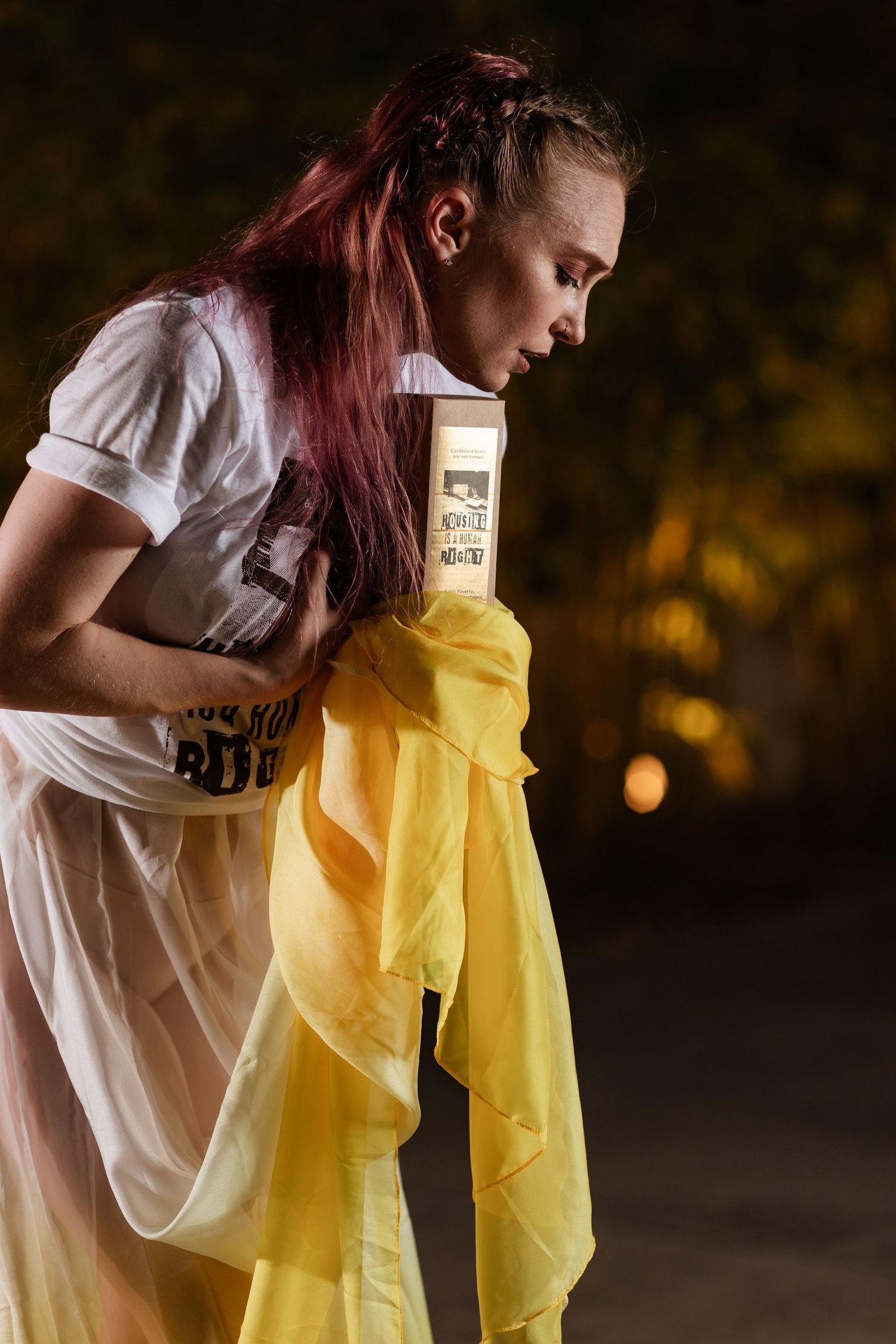 Young woman with red hair holding a glowing candle wrapped in yellow fabric outdoors at night.