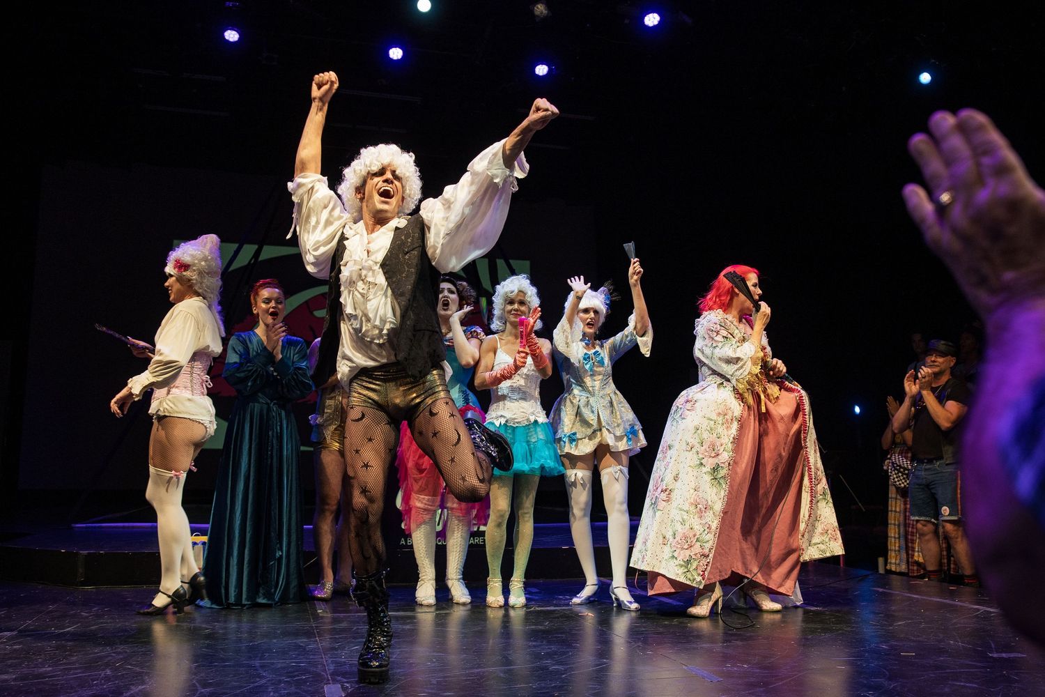 Burlesque ensemble in period costumes celebrate with raised fists during curtain call on darkened stage.