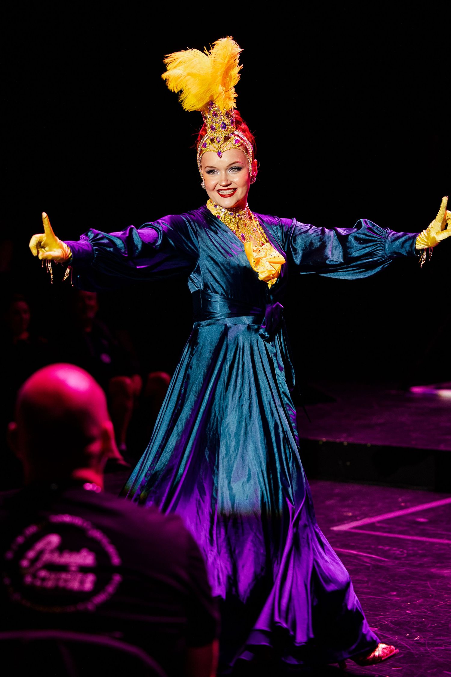 Smiling burlesque performer in blue-purple satin gown and gold feather headpiece spreads arms wide on stage.