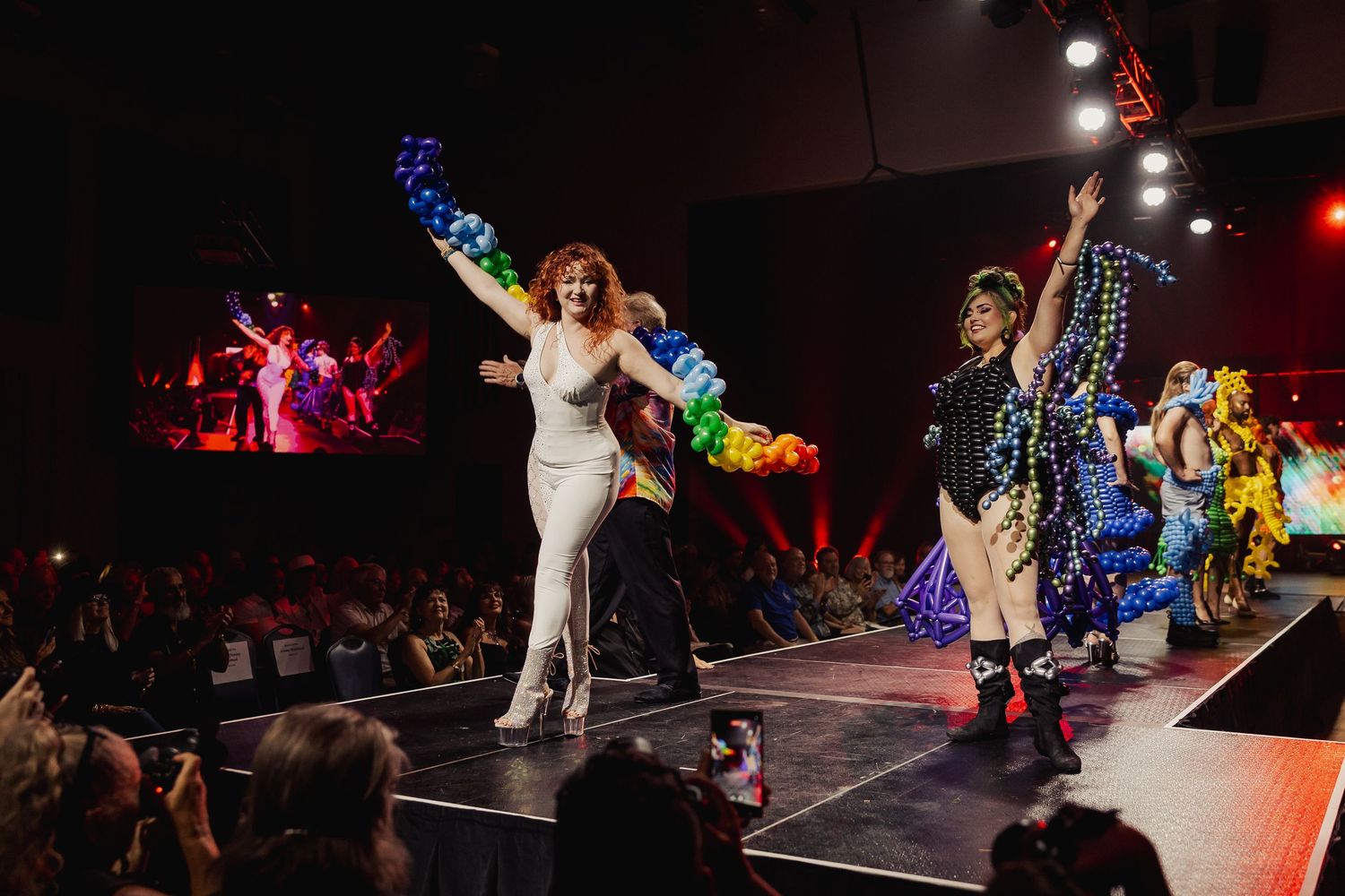 Models walk the finale runway at a wearable art fashion show with colorful balloon costumes.