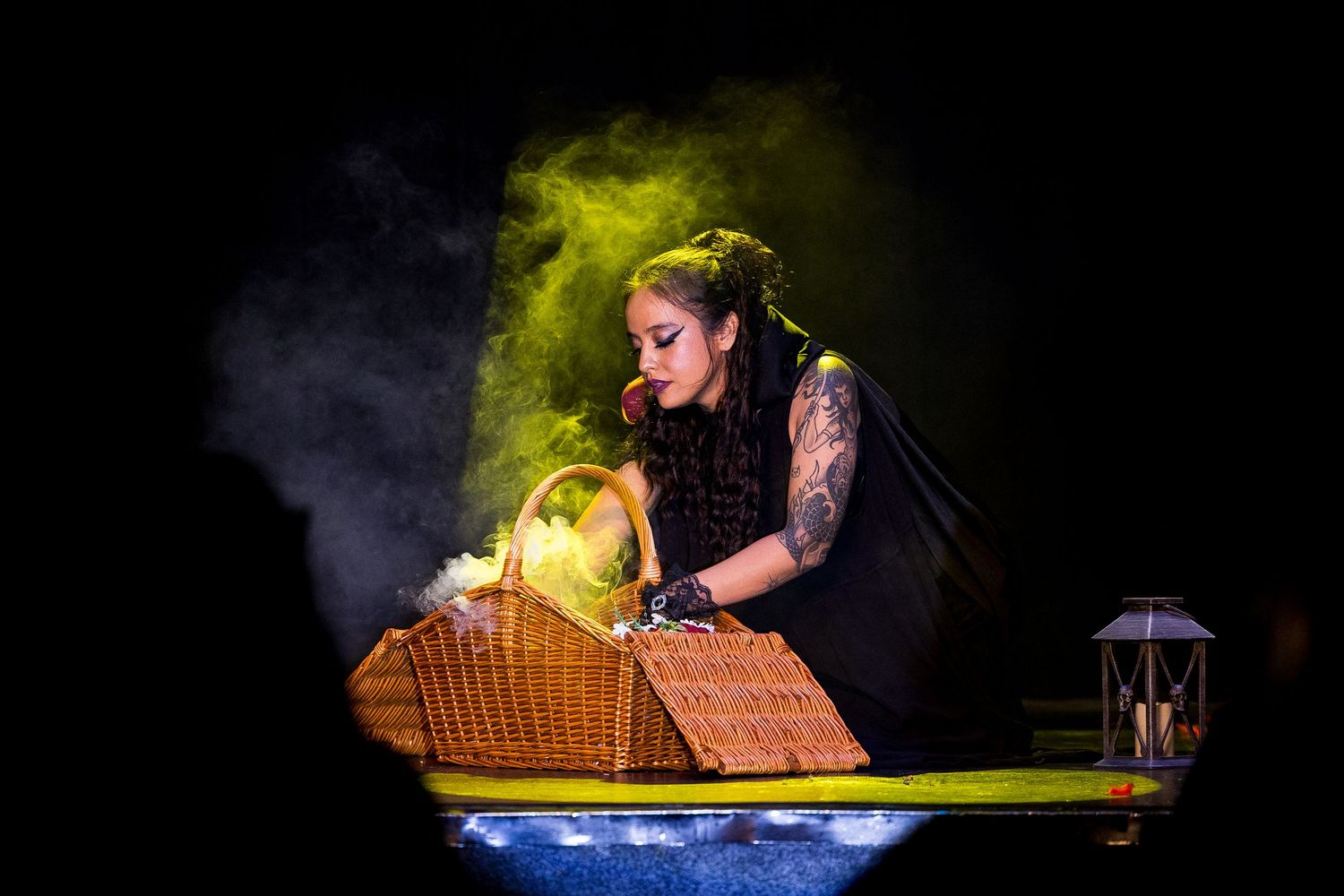 Tattooed performer in black costume leaning over wicker basket on dimly lit stage.