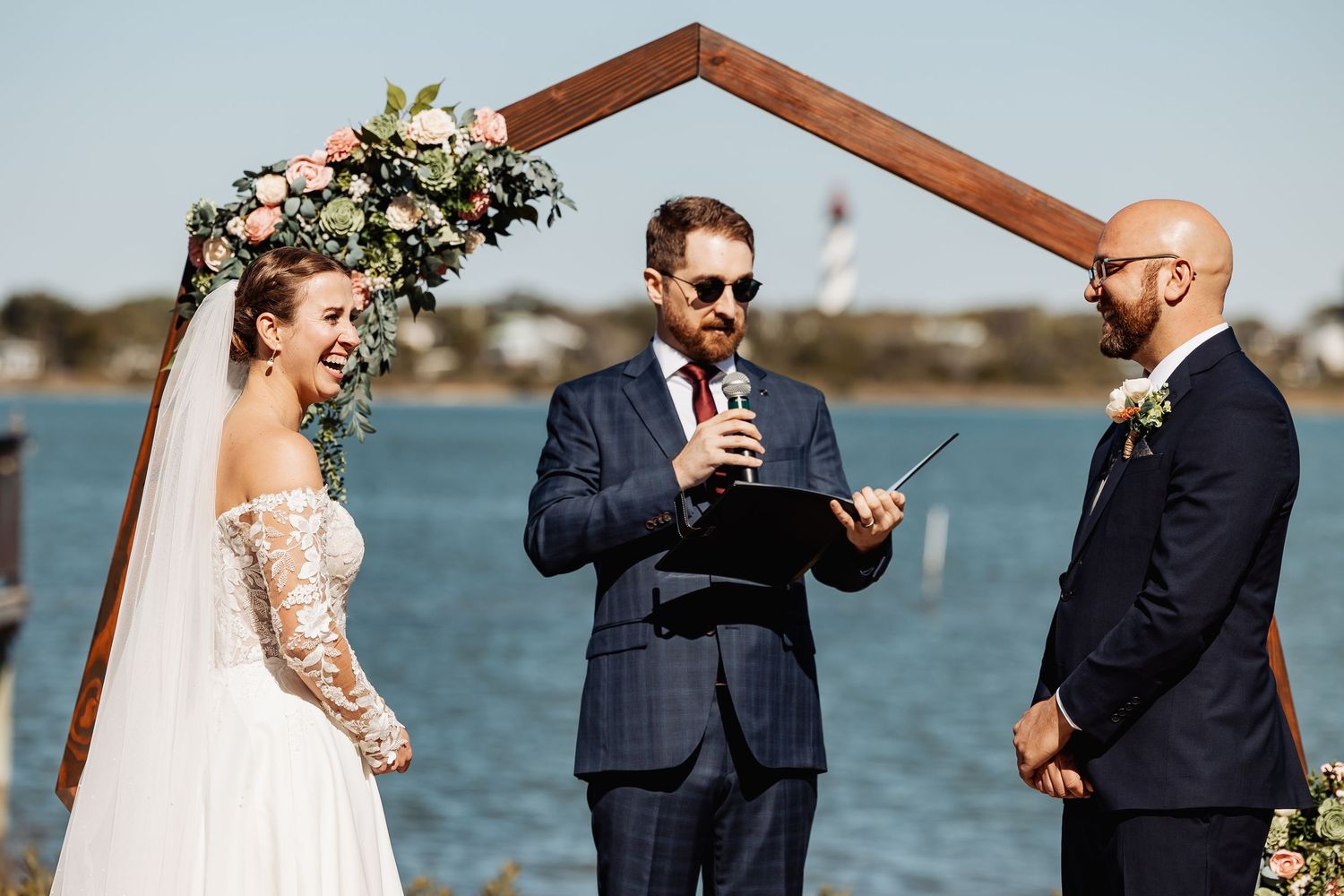 Bride laughs joyfully as officiant reads vows at outdoor waterfront ceremony with geometric wooden arch.