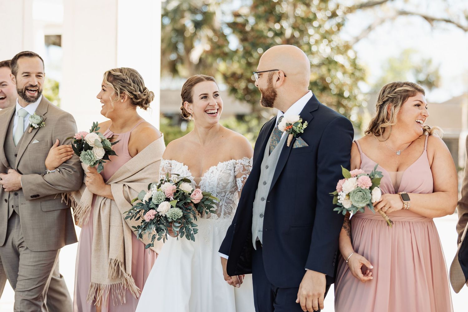 Bride laughing with wedding party, bridesmaids in pink dresses and groomsmen in tan suits.