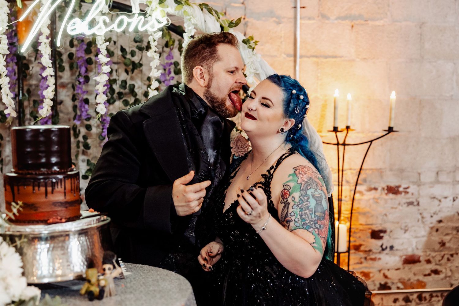 Newlyweds share a playful cake tasting moment beside their chocolate drip cake at gothic reception.