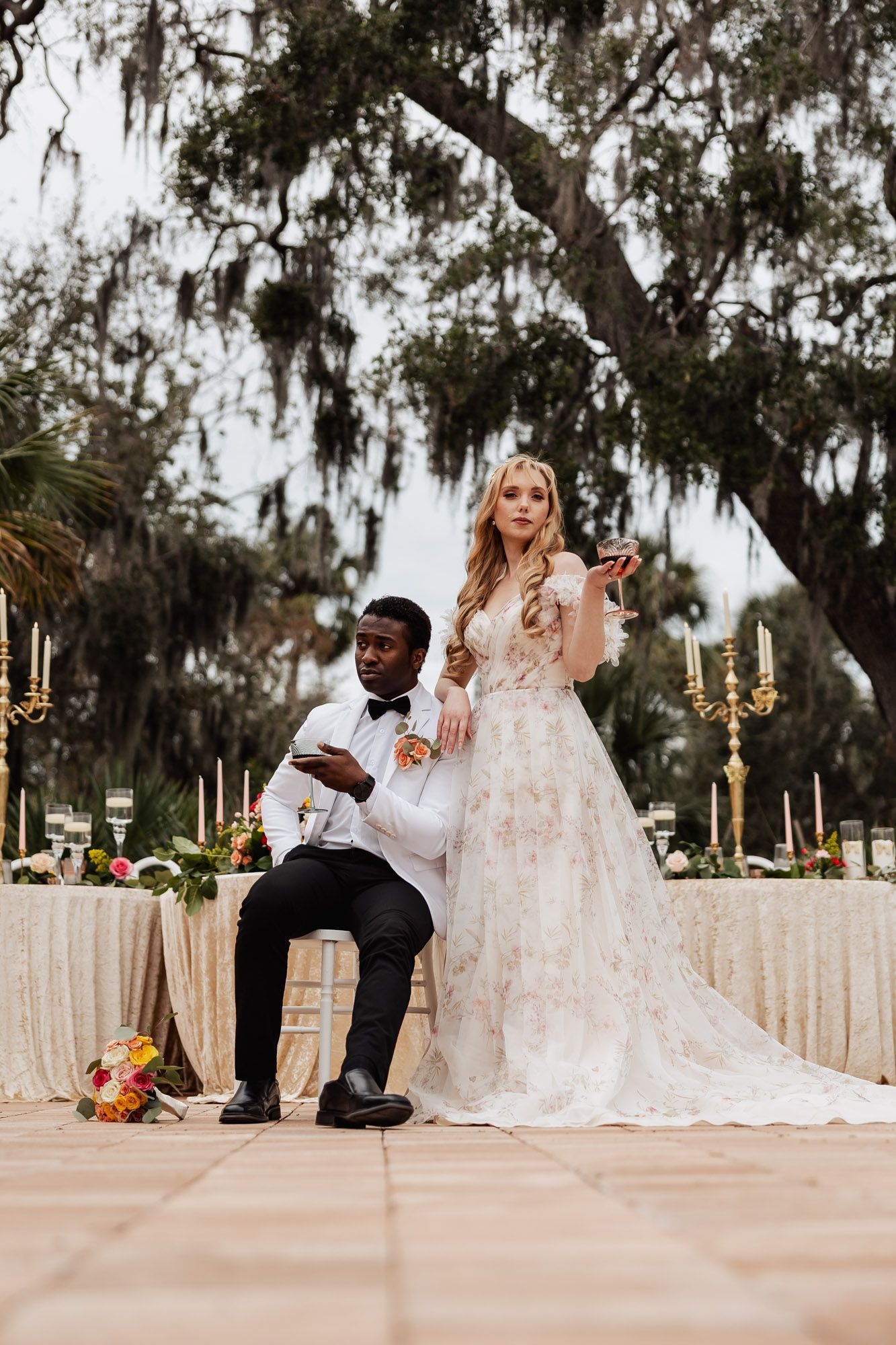 Interracial wedding couple holding wine glasses under Spanish moss trees at outdoor reception.