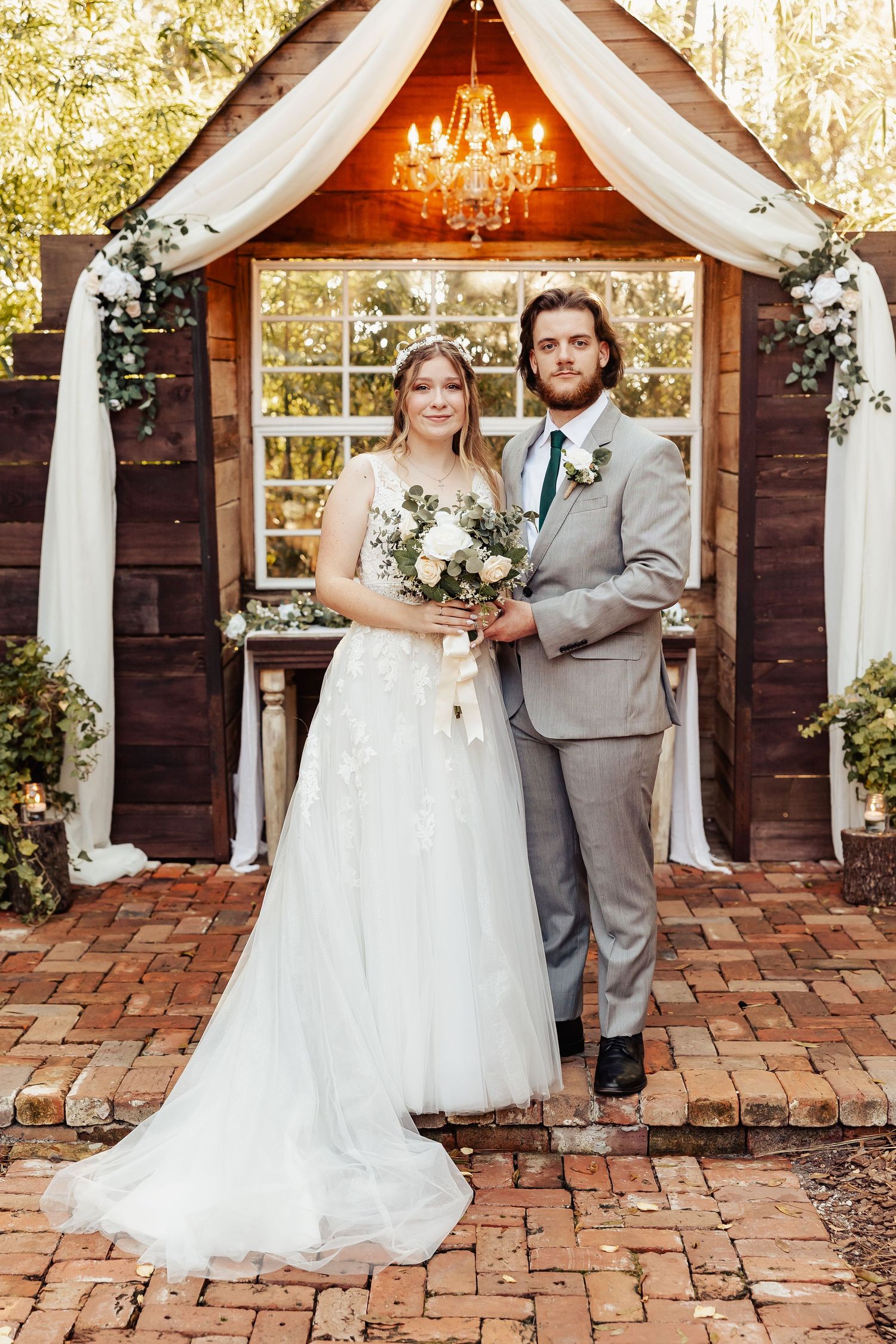 Wedding couple standing under decorated wooden arch with chandelier and white draping.