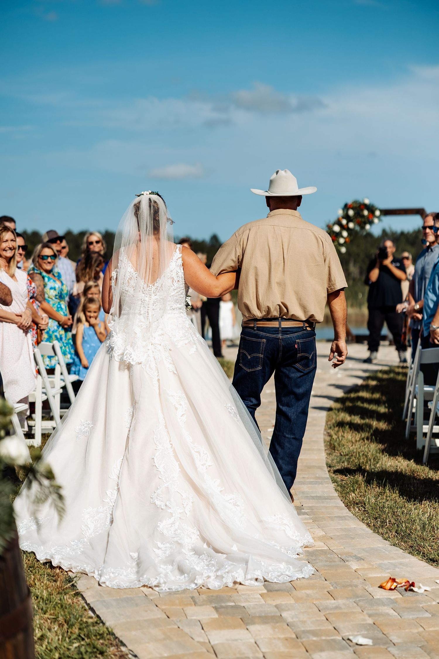 Outdoor wedding processional on stone pathway with white chairs and guests.