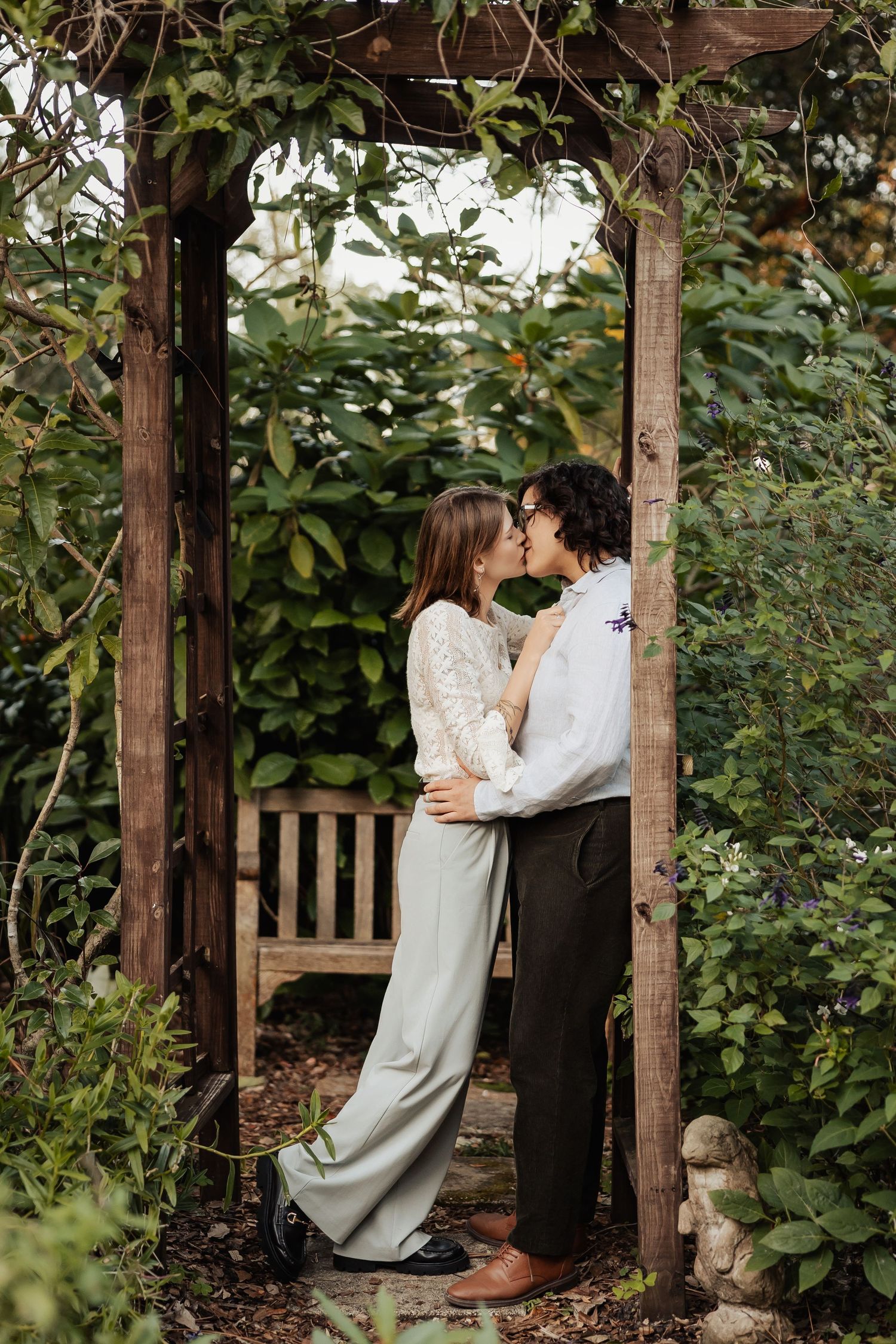 Two women share a kiss framed by a rustic wooden garden arbor covered in lush green vines.