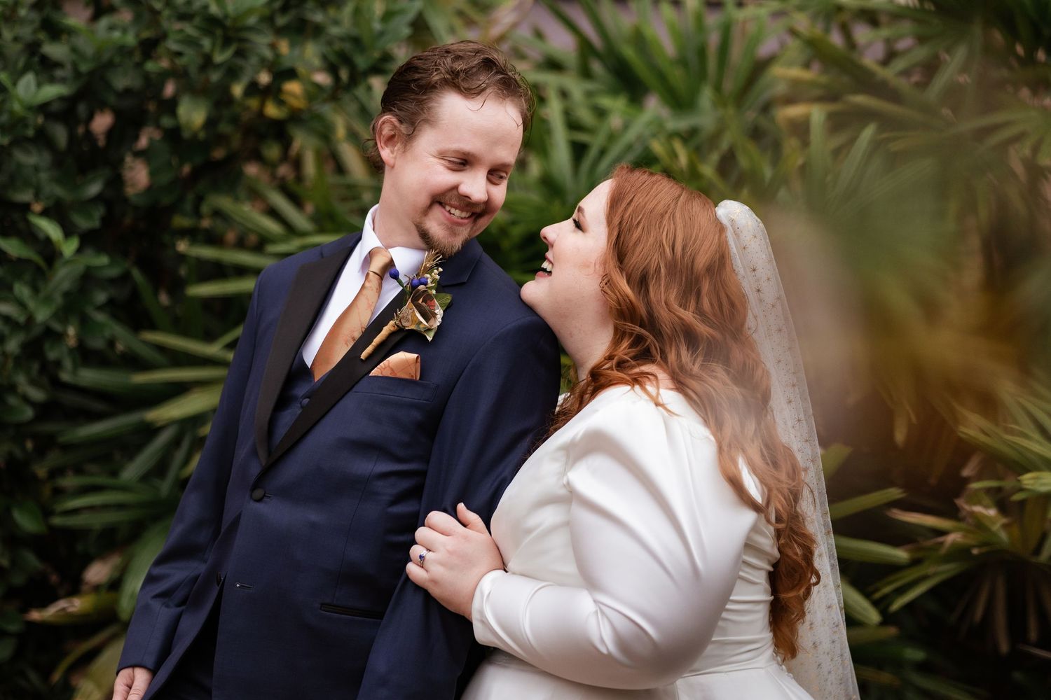 Smiling bride and groom gaze at each other surrounded by lush tropical greenery.