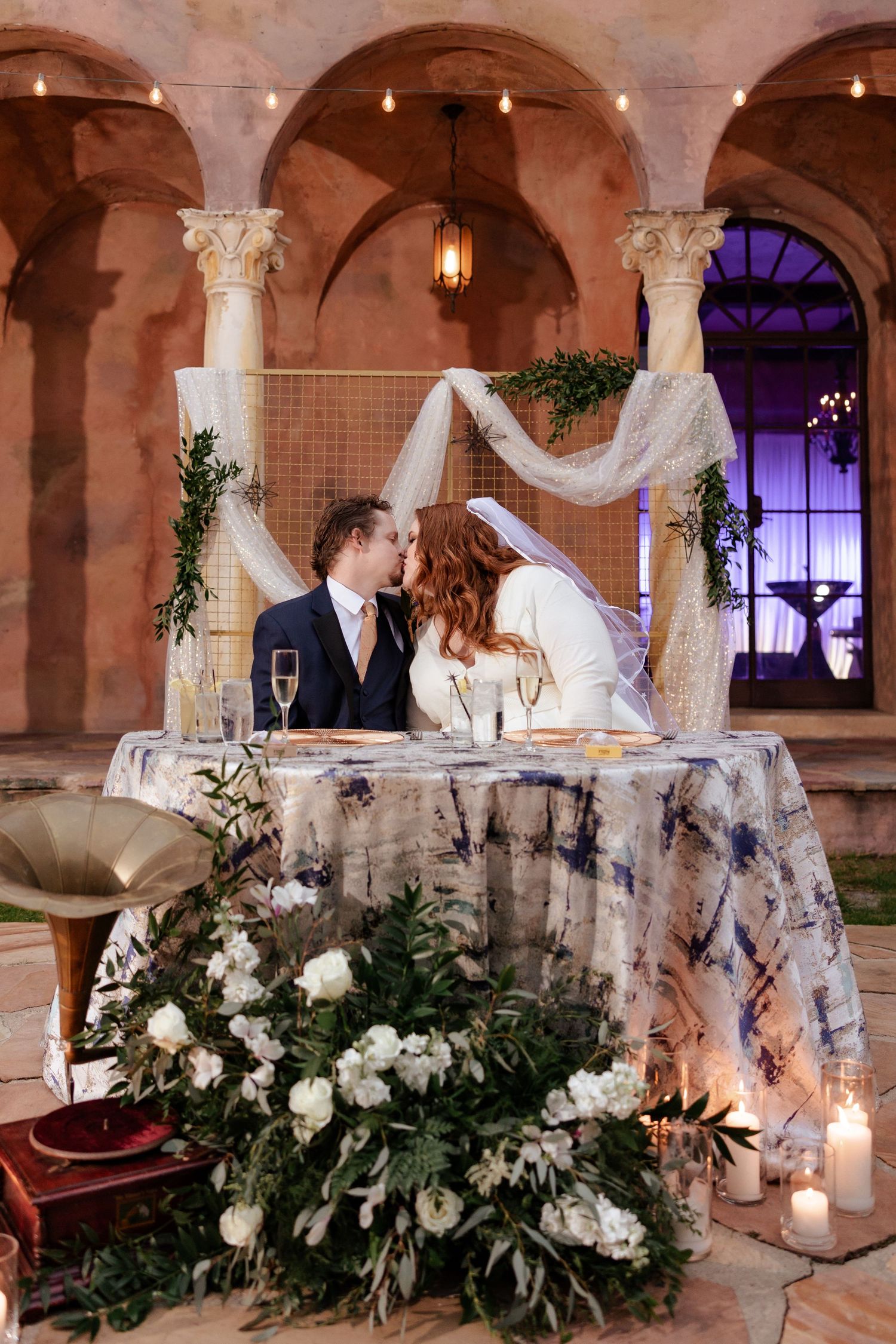 Bride and groom kissing at sweetheart table under floral arch at night.