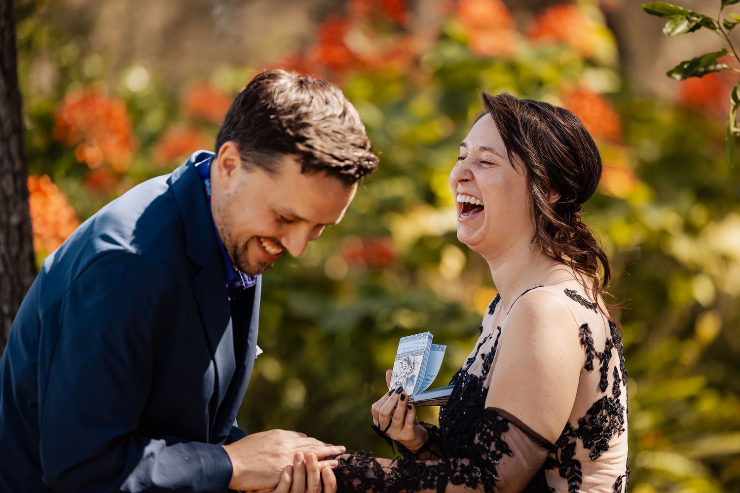Couple laughing together as groom places ring on bride's finger during outdoor wedding ceremony.