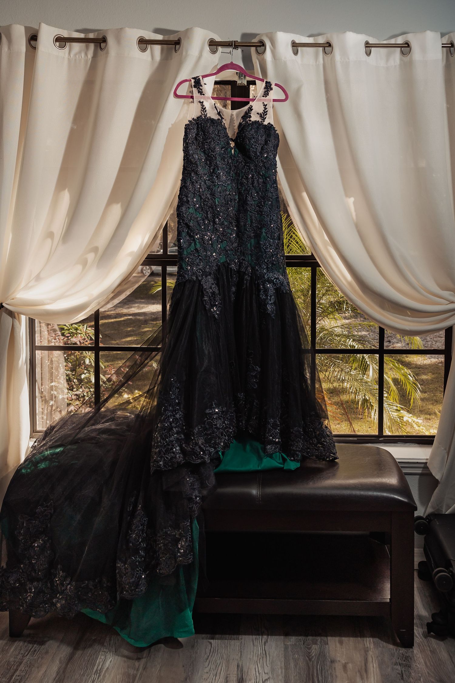 A black lace mermaid wedding gown hanging on a pink hanger in a sunlit window.