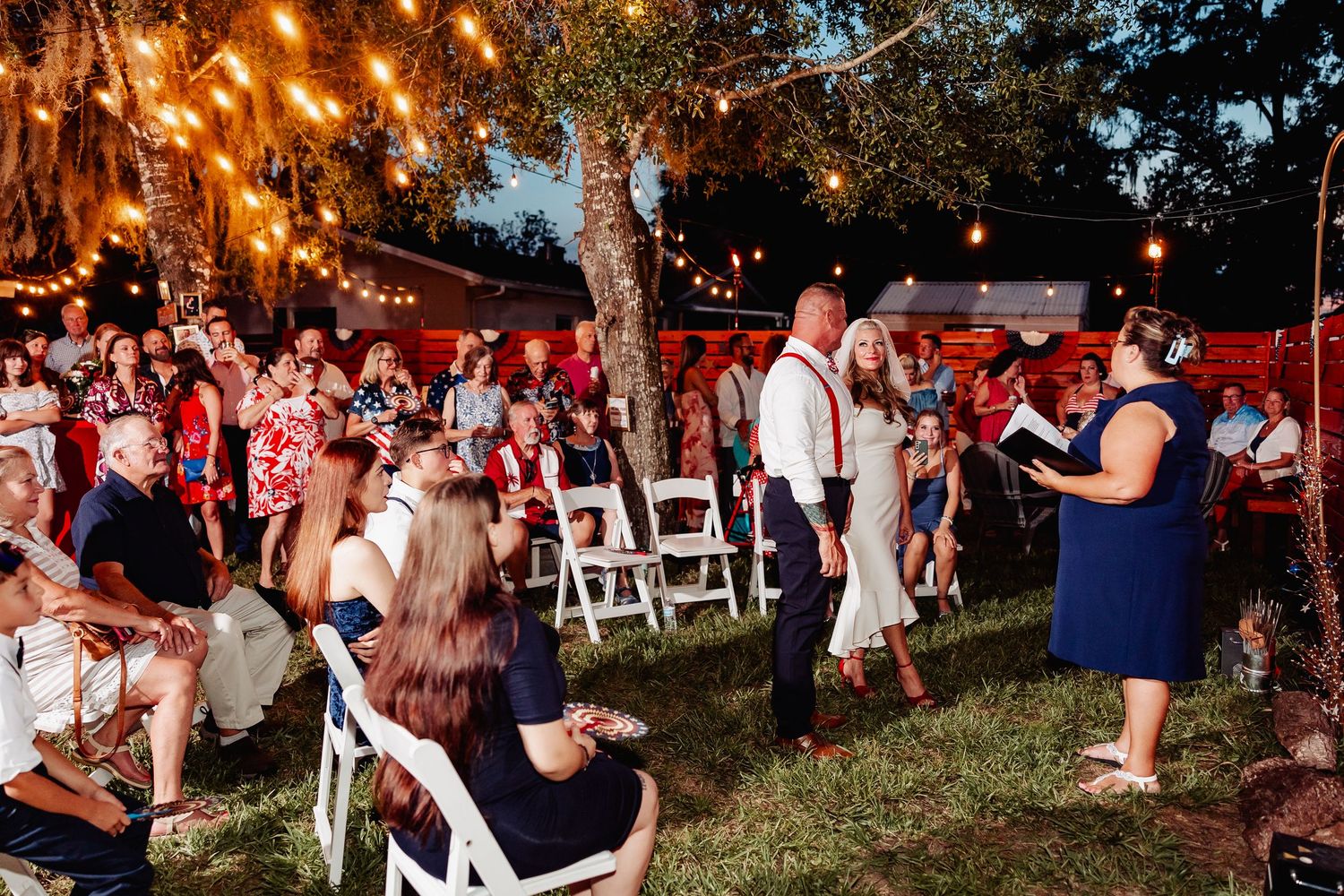 Outdoor backyard wedding ceremony at dusk with guests seated on white chairs under string lights in trees.