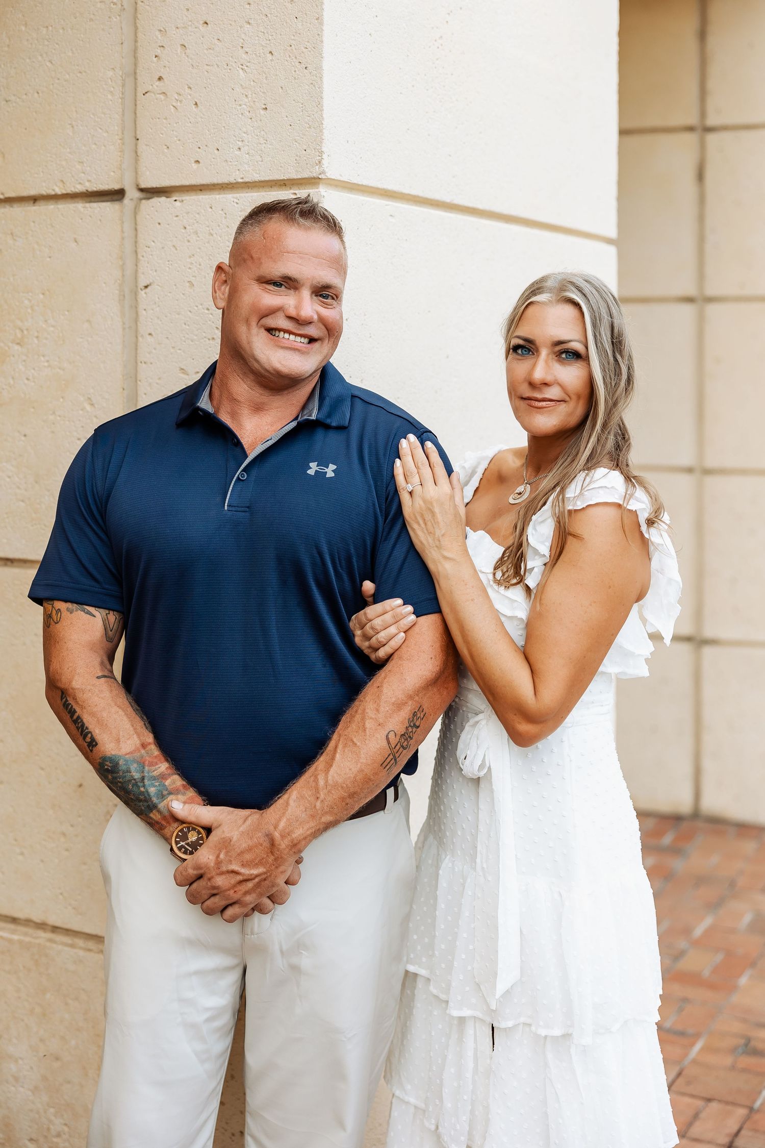 Smiling engaged couple poses near a stone column, woman in white dress displaying her engagement ring proudly.