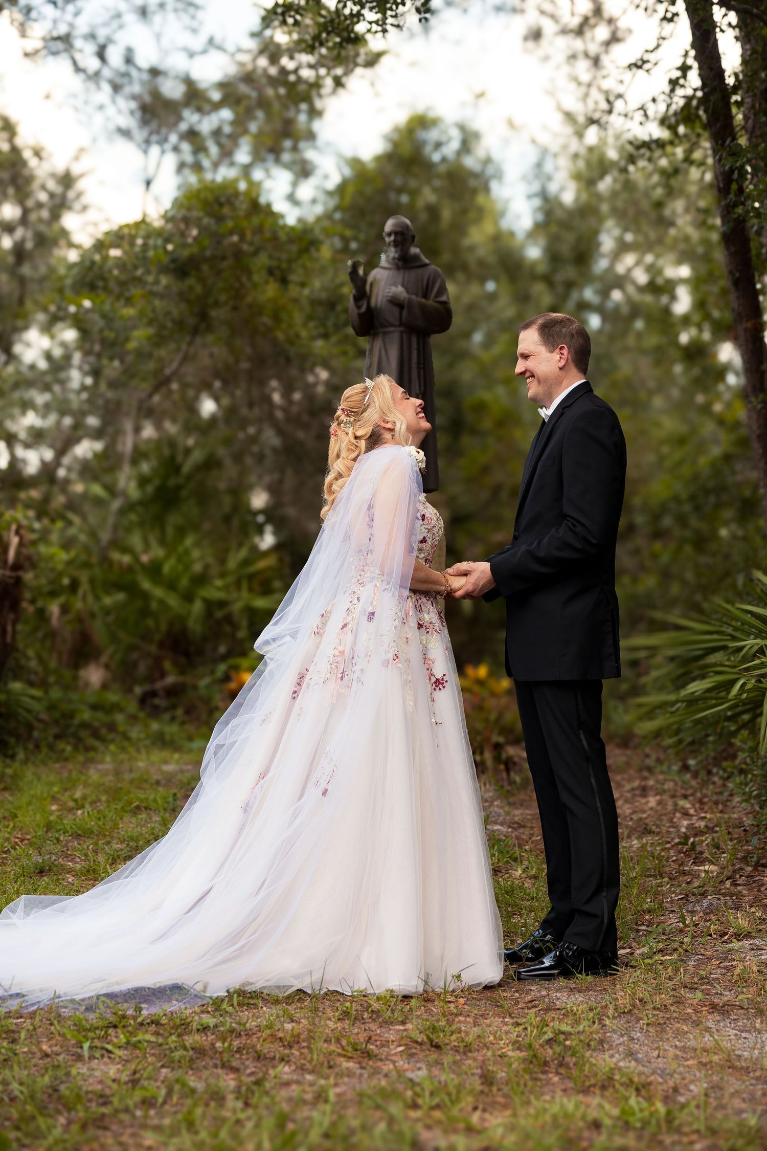 Bride and groom holding hands and laughing outdoors near a saint statue surrounded by greenery.