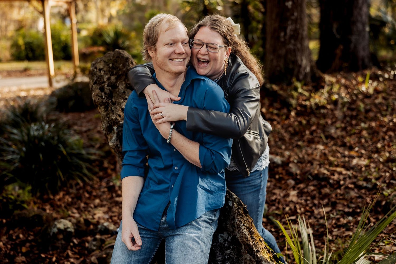 Laughing couple embracing playfully outdoors near a tree in a wooded park setting.