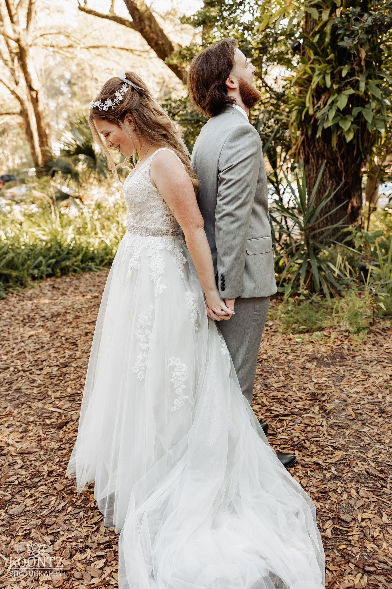 Bride in lace tulle gown and groom in gray suit holding hands back-to-back in a sunlit forest setting.