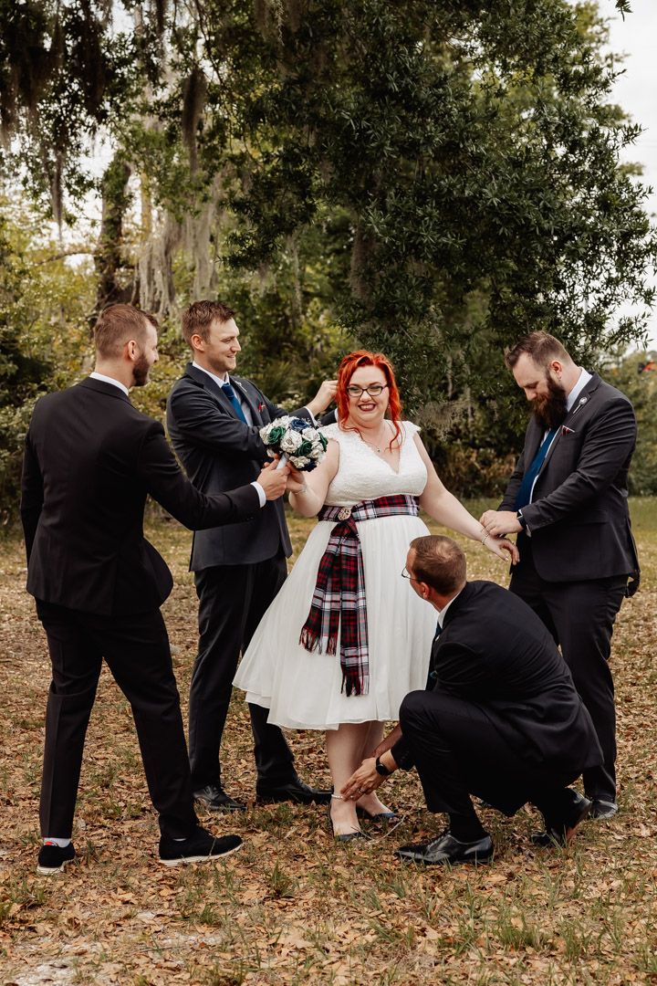 Groomsmen playfully attend to red-haired bride outdoors, fixing hair, holding bouquet, and tying shoe.