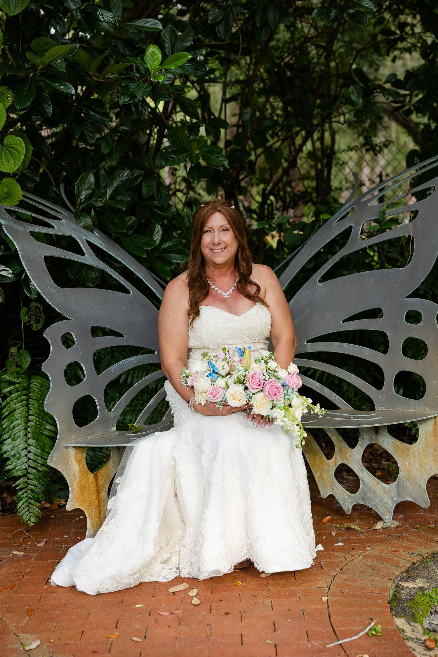 Radiant bride in lace gown sits on decorative butterfly bench holding colorful bouquet amid green foliage.