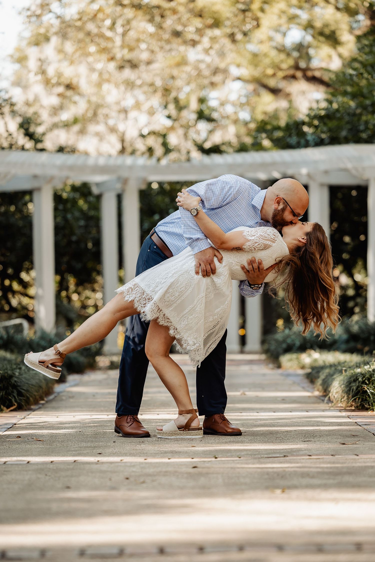Man dipping woman for a kiss on garden pathway with white pergola in background.