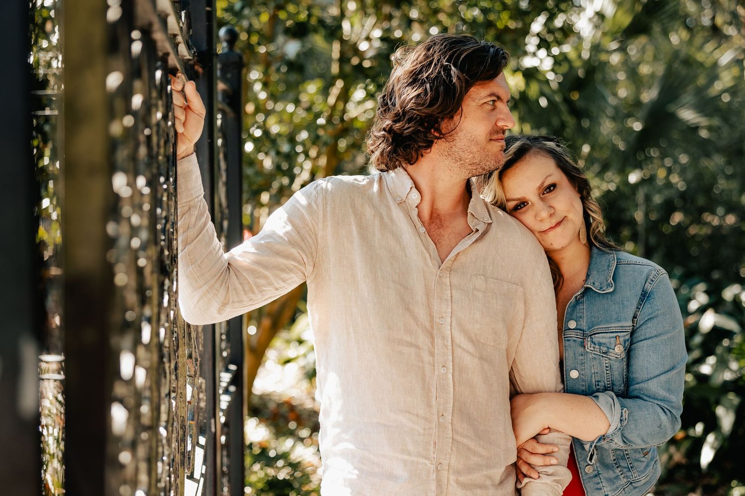 Couple standing by an ornate gate, woman in denim jacket resting her head on mans shoulder.
