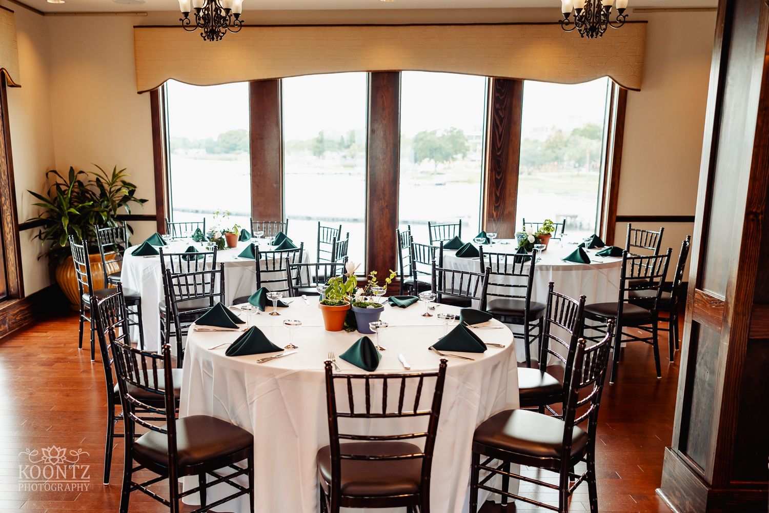 Elegant reception venue with round white-clothed tables and dark Chiavari chairs near large windows with natural light.