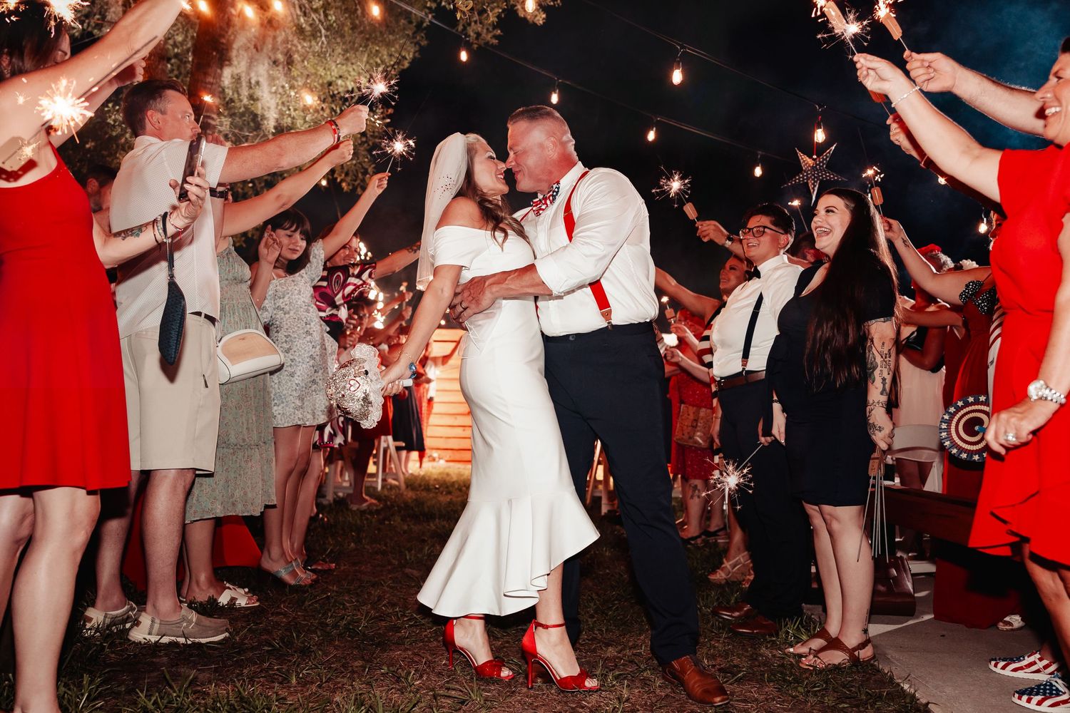 Wedding couple dances under sparklers at night surrounded by guests in red dresses.