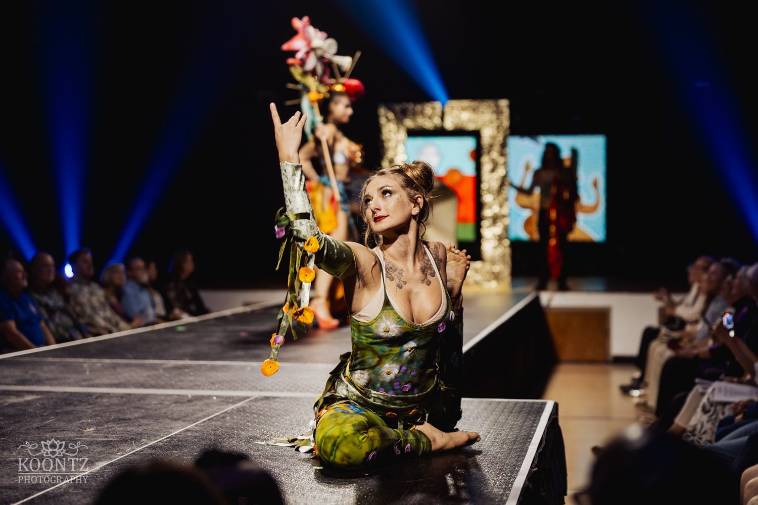 Fashion model poses dramatically on runway with flowers at fashion show with blue spotlights and audience visible.