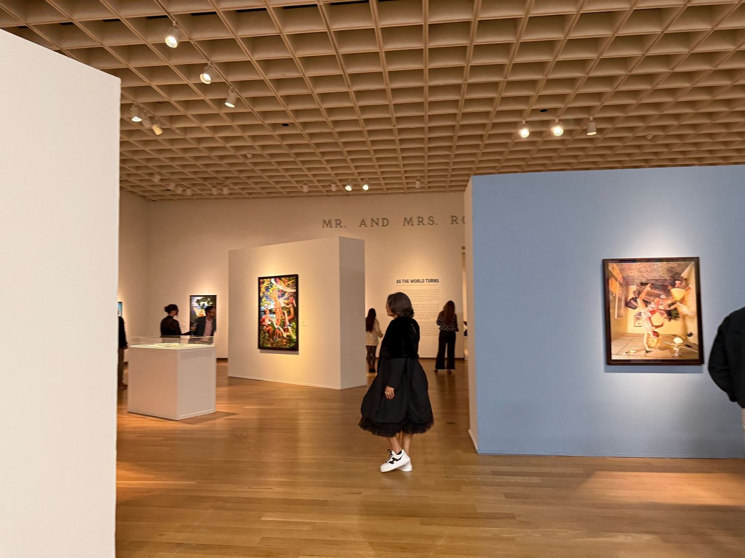 Art gallery interior with white walls displaying framed artwork under wooden lattice ceiling.