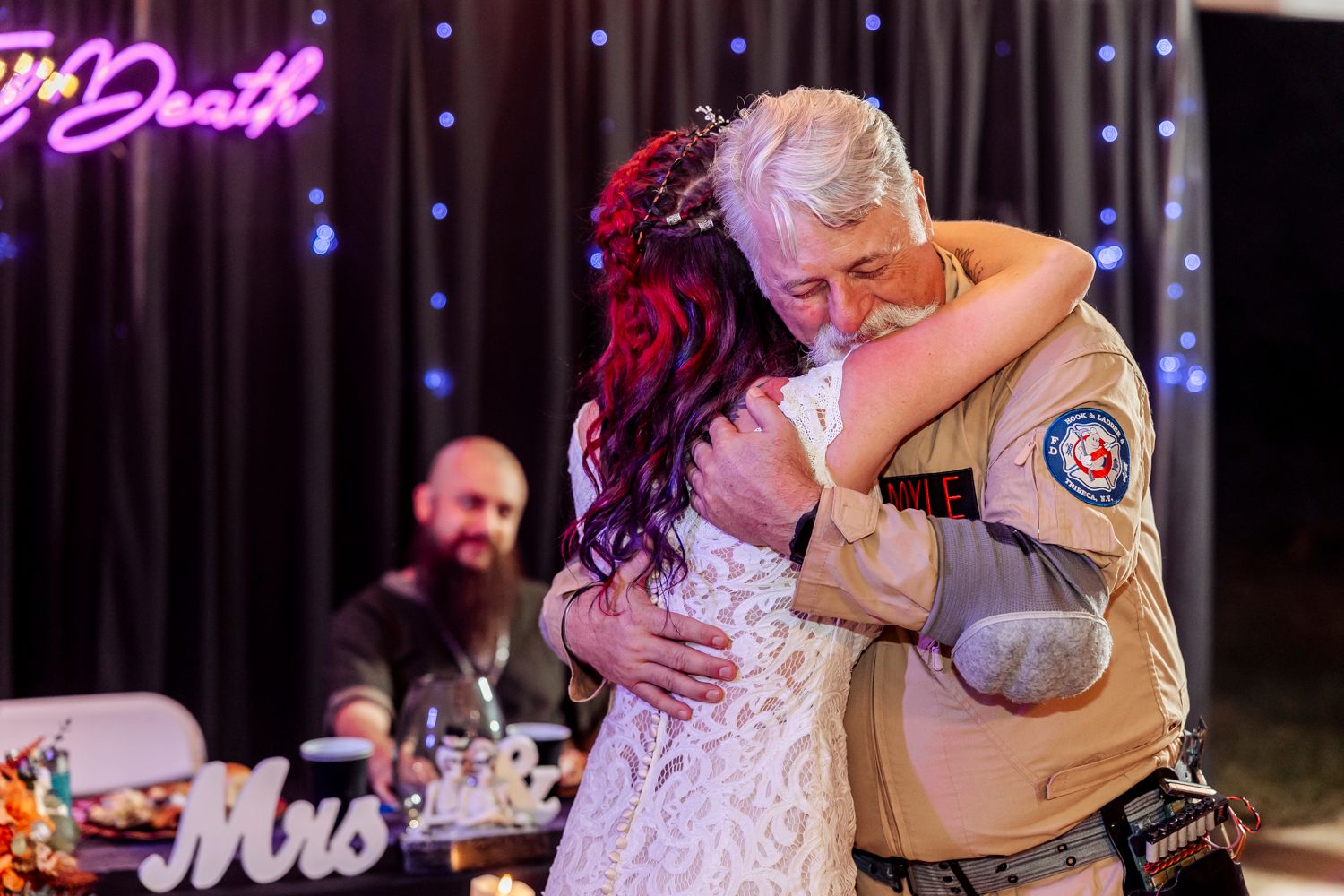 Couple shares an emotional dance moment at their wedding reception with purple neon sign glowing in background.