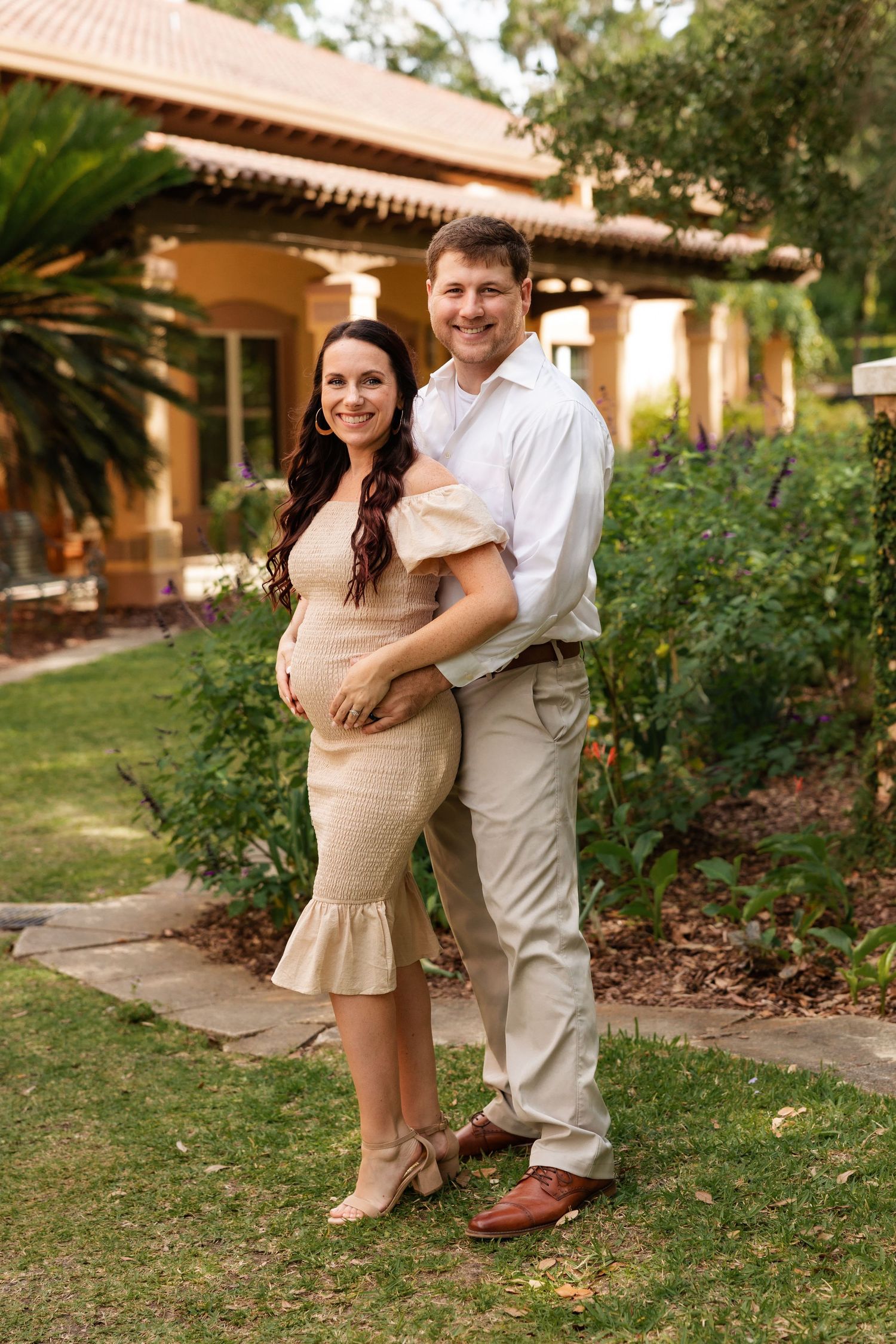 Couple posing in garden, pregnant woman in beige fitted dress beside partner.