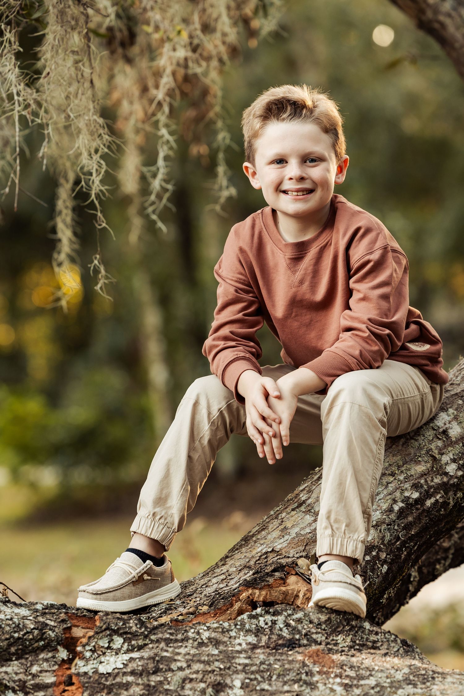 Smiling young boy in brown sweater and khaki pants sits on a mossy log in a sunlit forest.