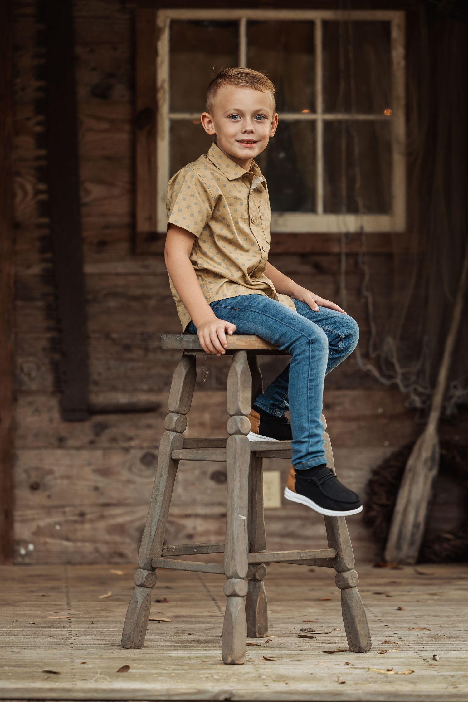 A young boy in a tan shirt and jeans sits on a rustic wooden stool in front of a vintage barn backdrop.