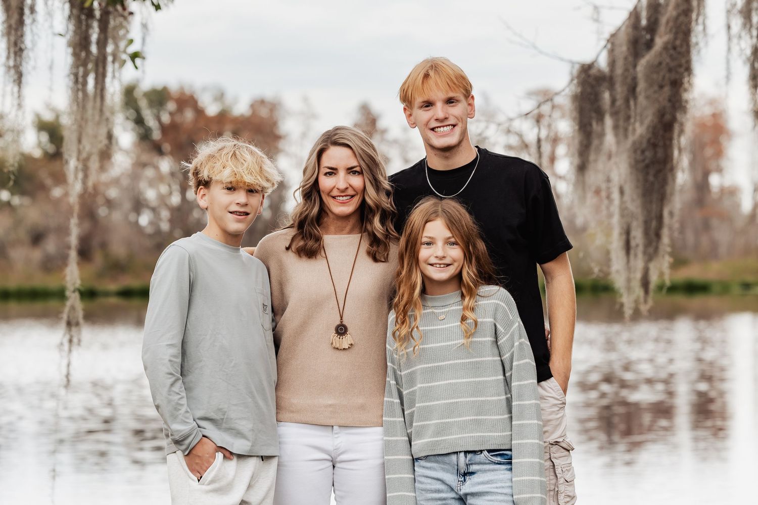 Family of four smiling together outdoors near a lake with Spanish moss trees in background.